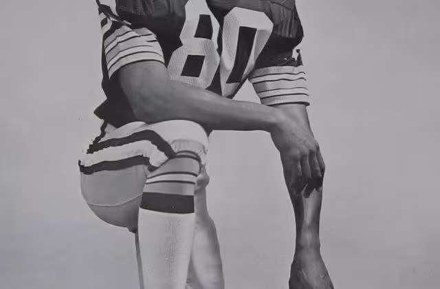 Black and white publicity photo of James Lofton on the Green Bay Packers, kneeling in Packers uniform, leaning on his GB helmet