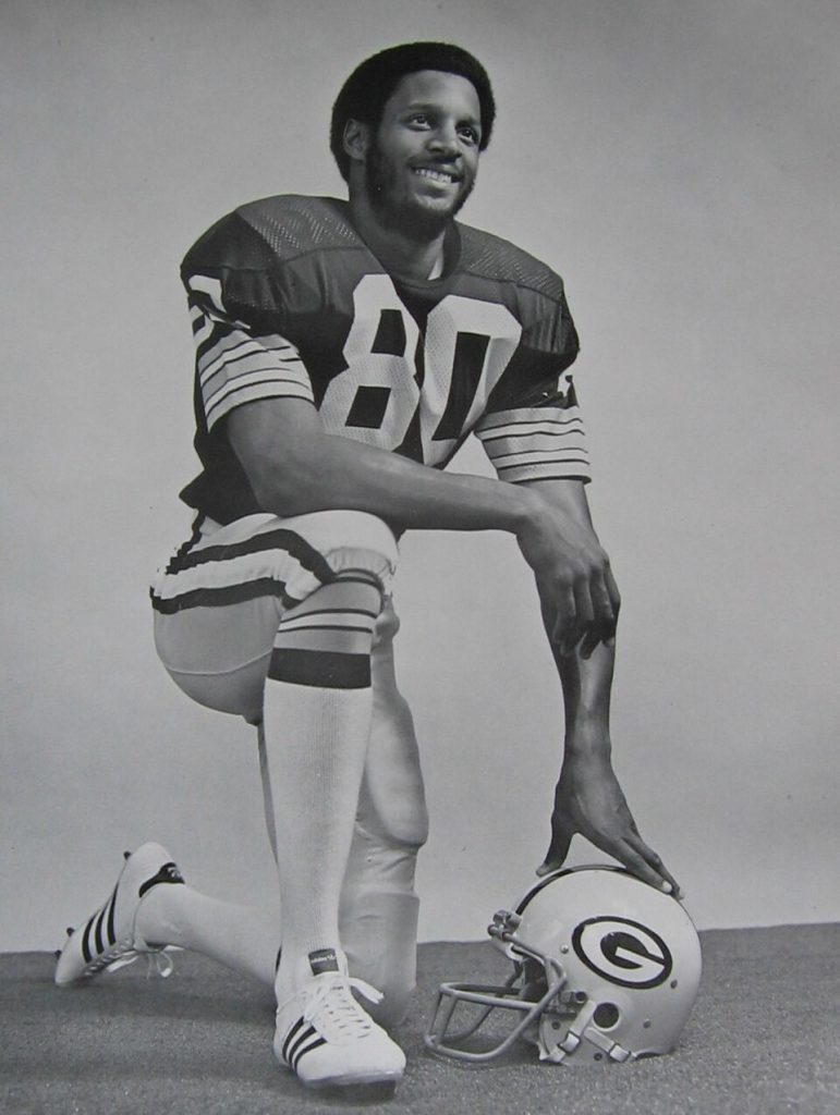 Black and white publicity photo of James Lofton on the Green Bay Packers, kneeling in Packers uniform, leaning on his GB helmet