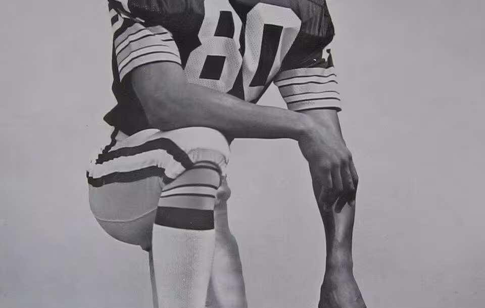 Black and white publicity photo of James Lofton on the Green Bay Packers, kneeling in Packers uniform, leaning on his GB helmet