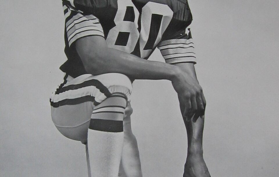 Black and white publicity photo of James Lofton on the Green Bay Packers, kneeling in Packers uniform, leaning on his GB helmet