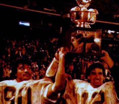 Blurred full color celebration photo of West Virginia Mountaineers Darryl Talley (#90) and Oliver Luck (#12) hoist the Peach Bowl trophy after their 1981 victory over the Florida Gators.