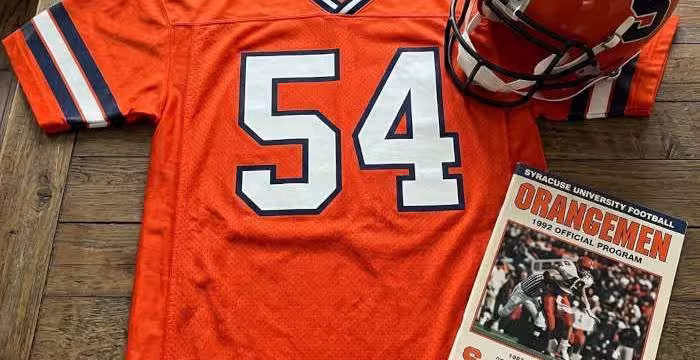 Gemini AI-created image of a 1992-era Syracuse American football jersey 54, without a name on it, and a matching period-correct helmet, the program cover appropriate to the era and team.