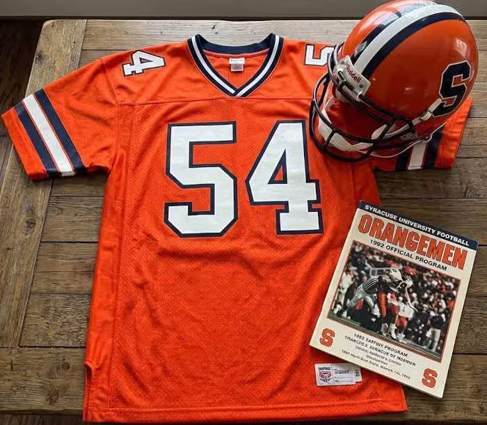 Gemini AI-created image of a 1992-era Syracuse American football jersey 54, without a name on it, and a matching period-correct helmet, the program cover appropriate to the era and team.