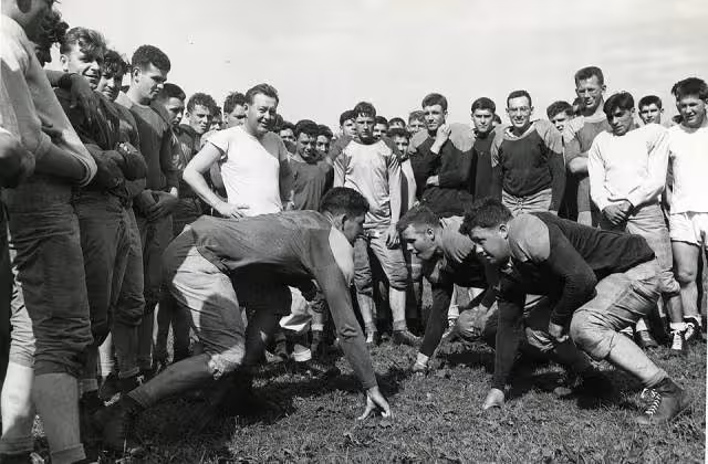 Black and white image photo of Football training has begun at constabulary headquarters at Bamberg, Germany. Practicing blocking are, Left to Right, Pfc, Bernard Clark of Sleepeye, Minn., and Pfc. Dang Davison of Downer's Grove, Ill. Coach Sandy Clark, wearing a white shirt, looks on from the rear left.