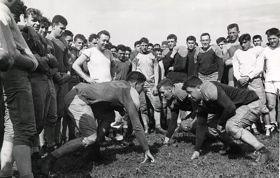 Black and white image photo of Football training has begun at constabulary headquarters at Bamberg, Germany. Practicing blocking are, Left to Right, Pfc, Bernard Clark of Sleepeye, Minn., and Pfc. Dang Davison of Downer's Grove, Ill. Coach Sandy Clark, wearing a white shirt, looks on from the rear left.
