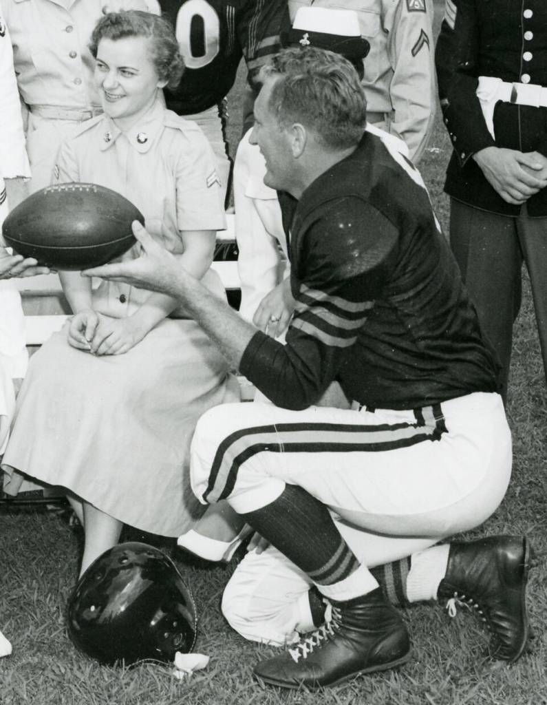 Black and white publicity photo of The original caption describes the photograph as: Military Personnel with Some of the Members of the Chicago Bears Football Team - In the group are George Blande, Cpl Jo Ruff, Cpl Sonia Schmit, Harry Jagade, fullback of the Bears, Larry Brink, and Pfc John Carbon.