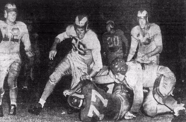 Game action photo of Vitamin Smith (bottom center, number 77) is tackled after a ten yard gain by Bob Levenhagen (bottom right) and Bobby Thompson (center, number 65) during an intrasquad game at Gilmore Stadium in Los Angeles, California on August 17, 1949.
