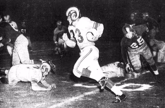 Black and white game action photo of Los Angeles Rams back Jerry Williams (center, with ball) returns a kickoff for 35 yards as Washington Redskins player Jim Canady (right, number 25) is in pursuit during a game on August 15, 1951 at Los Angeles Memorial Coliseum in Los Angeles, California.