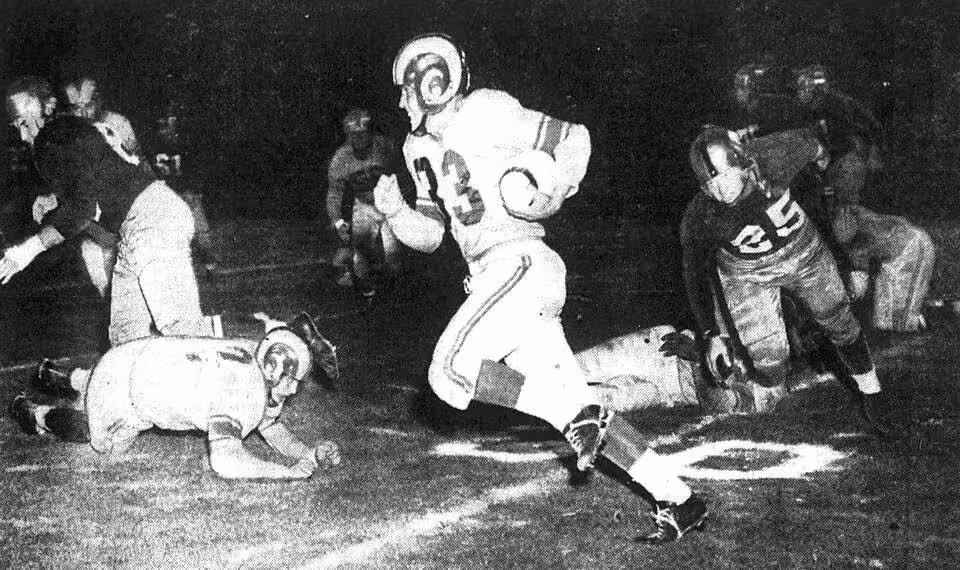 Black and white game action photo of Los Angeles Rams back Jerry Williams (center, with ball) returns a kickoff for 35 yards as Washington Redskins player Jim Canady (right, number 25) is in pursuit during a game on August 15, 1951 at Los Angeles Memorial Coliseum in Los Angeles, California.