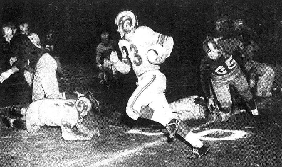 Black and white game action photo of Los Angeles Rams back Jerry Williams (center, with ball) returns a kickoff for 35 yards as Washington Redskins player Jim Canady (right, number 25) is in pursuit during a game on August 15, 1951 at Los Angeles Memorial Coliseum in Los Angeles, California.