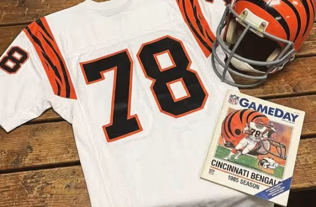 Gemini AI-created mage of a 1985-era Cincinnati NFL American white football jersey 78, without a name on it, and a matching period-correct helmet, the program cover appropriate to the era and team.