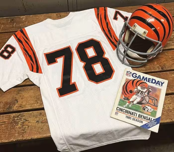 Gemini AI-created mage of a 1985-era Cincinnati NFL American white football jersey 78, without a name on it, and a matching period-correct helmet, the program cover appropriate to the era and team.