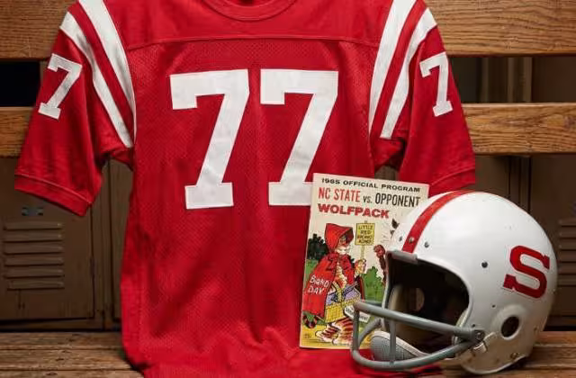 Gemini AI-created image of a 1965-era NC State American football jersey 77, without a name on it, and a matching period-correct helmet, the program cover appropriate to the era and team.