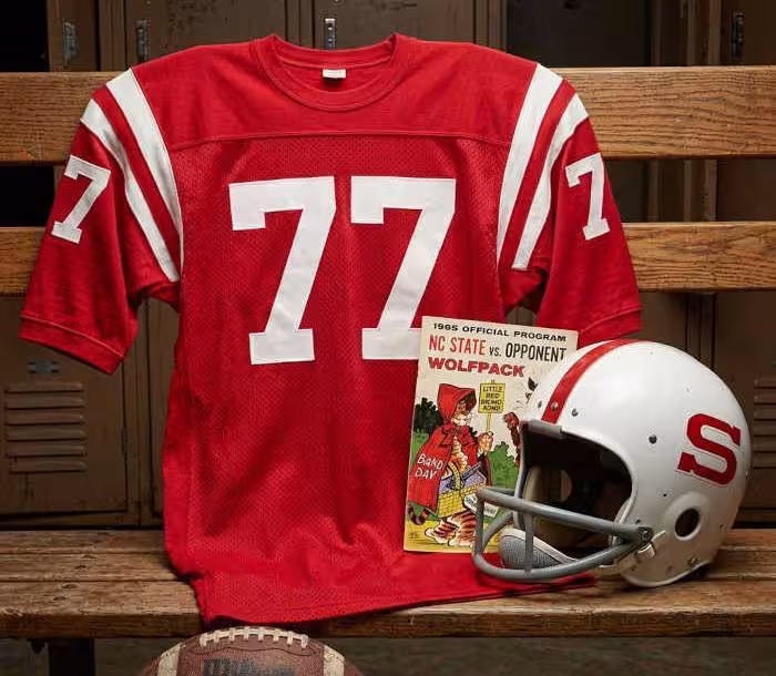 Gemini AI-created image of a 1965-era NC State American football jersey 77, without a name on it, and a matching period-correct helmet, the program cover appropriate to the era and team.