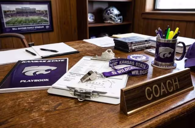 Gemini AI-created image of a wooden desk on an American college football coaches office with a placard that says "Coach" a clipboard, a playbook, a whistle and lanyard, and some purple items that refer to Kansas State football.